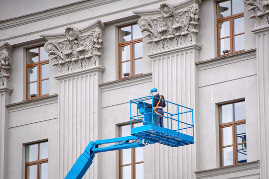 Construction worker in lift bucket of crane restore and repair historic facade of building. Man in cradle, restoring plaster decoration on facade. Workers painting building, external building work