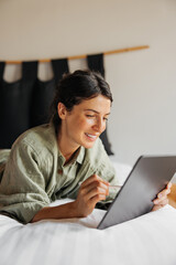 Smiling young caucasian girl signs electronic document on tablet, lies on bed. Brunette uses wireless device. Education, business, work from home concept.