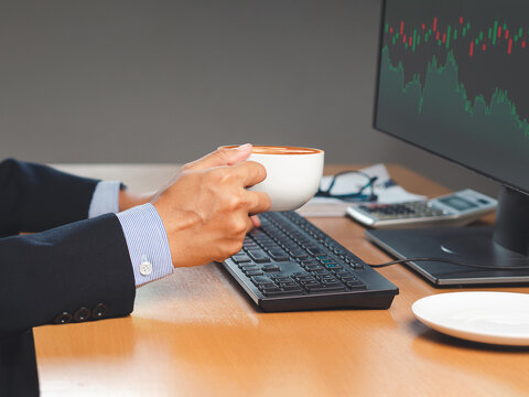 Hand of a businessman holding a coffee cup and looking at a graph stock on a screen monitor while sitting in the office