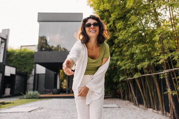 Smiling young caucasian brunette woman reaches for camera with her hand standing outdoors. Girl in good mood wears casual clothes. Happy weekend concept