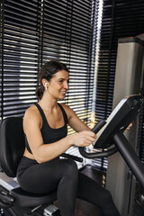 Fototapeta na wymiar Beautiful caucasian young brunette woman doing cardio on exercise bike in gym. Girl wears black top and leggings in training. Healthy lifestyle concept