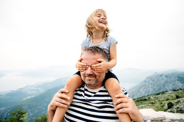 dad and daughter smiling and having fun together. daughter sitting on dad's shoulders and covers dad's eyes with hands. They are standing on the view point on Kotor bay, Montenegro. Wide angle photo