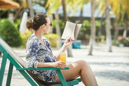 Happy smiling woman sitting in deck chair reading book at beach  in resort.