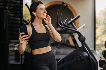 Happy young caucasian woman using mobile phone listening to music after fitness workout. Brunette wearing black top and leggings looks away. Relaxation, healthy lifestyle concept
