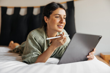 Enthusiastic young caucasian woman using stylus signs digital tablet lying on bed. Brunette wears...