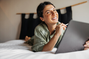 Happy young caucasian woman is using graphics tablet with stylus lying on bed. Brunette looking at camera spending leisure time at home. Hobby concept