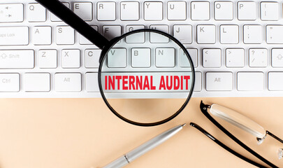 Text INTERNAL AUDIT on keyboard with magnifier , glasses and pen on beige background