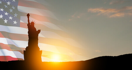 Statue of Liberty with a large american flag and sunset sky on background.