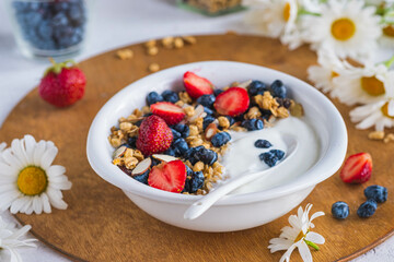 Unsweetened yogurt with granola, strawberries, honeysuckle and almond petals in a white bowl on a wooden stand on a light concrete background. Quick breakfast recipes, granola.