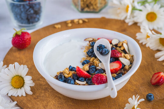 Unsweetened yogurt with granola, strawberries, honeysuckle and almond petals in a white bowl on a wooden stand on a light concrete background. Quick breakfast recipes, granola.