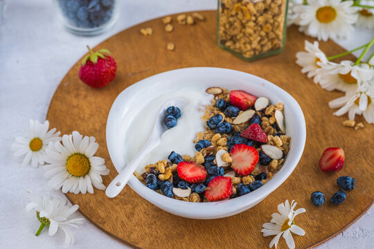 Unsweetened yogurt with granola, strawberries, honeysuckle and almond petals in a white bowl on a wooden stand on a light concrete background. Quick breakfast recipes, granola.