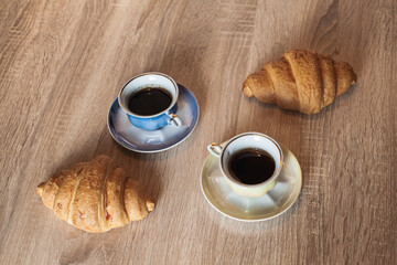 A couple of cups of hot fragrant black coffee in a saucer with croissants on a light brown wooden table.