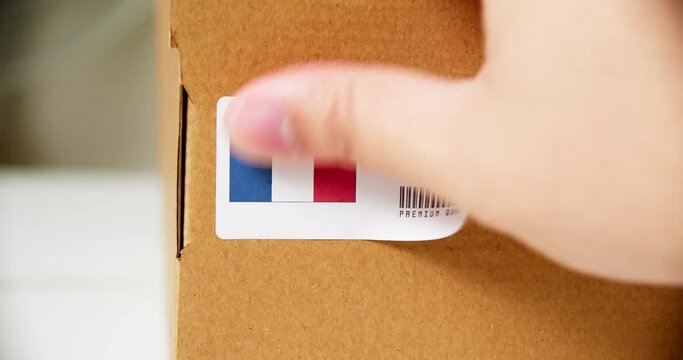 Hands applying MADE IN FRANCE flag label on a shipping cardboard box with products. Close up shot with premium quality barcode.