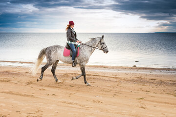 dressing jeans, jacket and spring hat horsewoman rides astride a dappled horse along seaboard