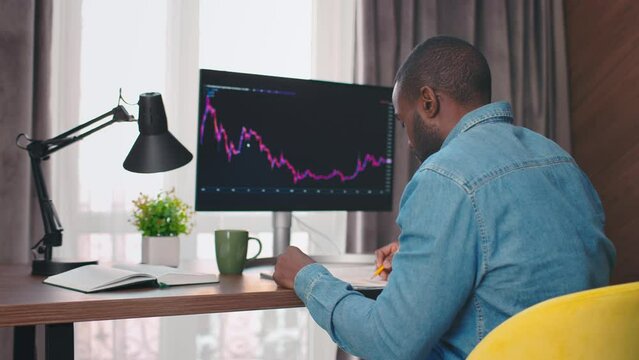 Side view close up shot of african american broker writing in notepad, checking computer with graphs and diagrams on sceen. Crypto currency trader. 4k. Office work concept.