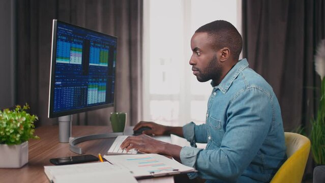 Side view close up portrait of african american trader typing and looking at computer with graphs and tables on sceen. Trader cheсking stock graphs, stock market. 4k. Office work concept.