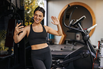 Fototapeta na wymiar Pretty young caucasian woman communicates by video link via smartphone standing in gym. Brunette wears sporty black top and leggings. Technology concept