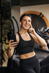 Smiling caucasian young woman listens to music through wireless headphones on smartphone in gym. Brunette wears sporty black top and leggings. Keep yourself in shape concept.