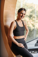 Fototapeta na wymiar Pretty caucasian young woman looks at camera, stands in room at window. Brunette wears sports top and leggings. Healthy lifestyle concept