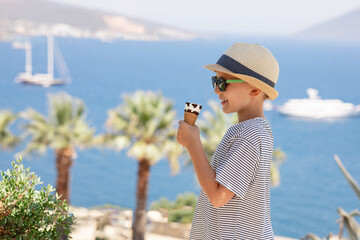 Young boy on summer vacation. Portrait of happy child with sea on background. Handsome smiling kid in straw hat and sunglasses eating ice cream outdoor.