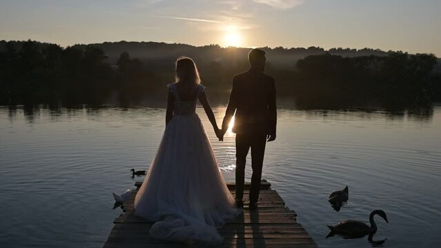 The newlyweds walk together hand in hand towards the lake at sunset