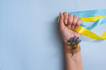 Fototapeta premium Female hand with yellow and blue ribbons and flowers on a blue background. Love and pray to Ukraine concept