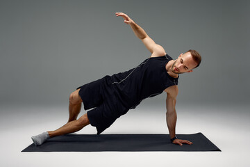 Confident young fitness man exercising on a fitness mat indoors, over gray wall background