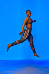 Obraz premium Happy african american woman wearing sportive clothes showing plus size hips. Body positive concept. Blue light on background