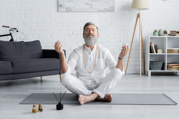 bearded yoga master meditating in easy pose near aroma sticks and scented oils.