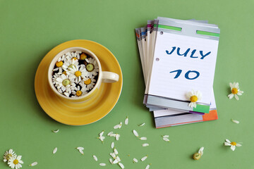 July 10 calendar: numbers 10 and the name of the month july on a sheet calendar, next to a cup of chamomile tea, pastel background, top view.