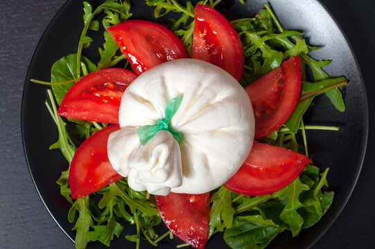 on a black plate is a salad of spinach and arugula greens, sliced tomatoes and Italian burrata cheese on a black background, top view, close-up.