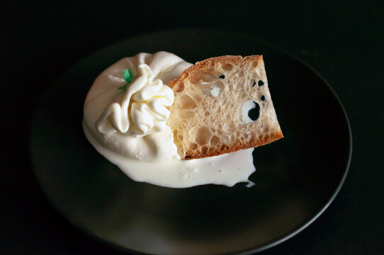 the cut Italian burrata cheese lies on a black plate, cream flows out of the burrata, a slice of porous bread is placed in the cut, side view.