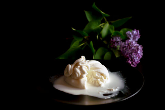 the cut Italian burrata cheese lies on a black plate, cream flows out of the envelope of the burrata. in the background lies a branch of blooming purple lilac. copy space.