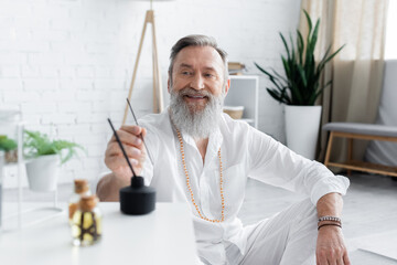 happy healing master holding aroma stick near scented oils on blurred foreground.