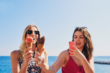 Two friends taking photos of ice cream eating on beach using smart phone technology on adventure...