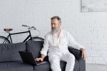 senior mastermind coach in white clothes using laptop while sitting on sofa at home.
