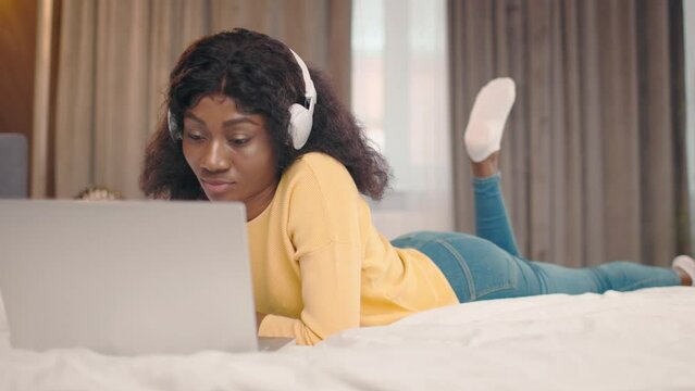 Beautiful young african american woman in headphones using laptop typing on keyboard and lying in comfy bed alone and smiling. People and lifestyle concept.