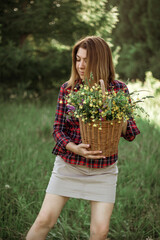 Summer lifestyle portrait of a beautiful young woman smiling and holding a basket with a bouquet of wild flowers. The concept of happiness and love