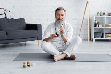 bearded healing guru sitting in easy pose with aroma sticks near bottles with flavored oils.