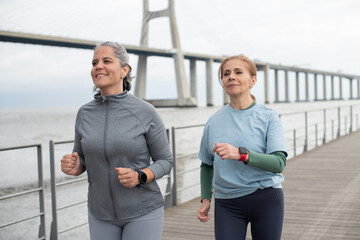 Happy senior friends jogging on embankment. Women in sportive clothes exercising on cloudy day. Sport, friendship concept