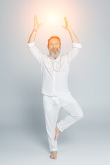 full length of yoga guru meditating in tree pose near shining chakra on grey.