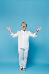 barefoot bearded man with closed eyes meditating in tree pose with open arms on blue.