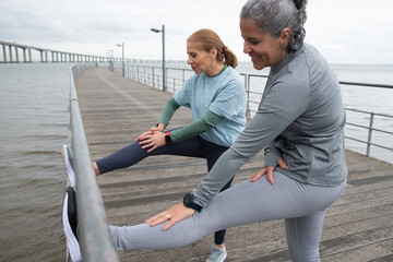 Lively senior friends exercising on embankment. Women in sportive clothes stretching on cloudy day. Sport, friendship concept