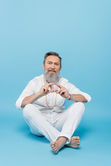 senior guru tutor showing hakini mudra while sitting with crossed legs on blue.