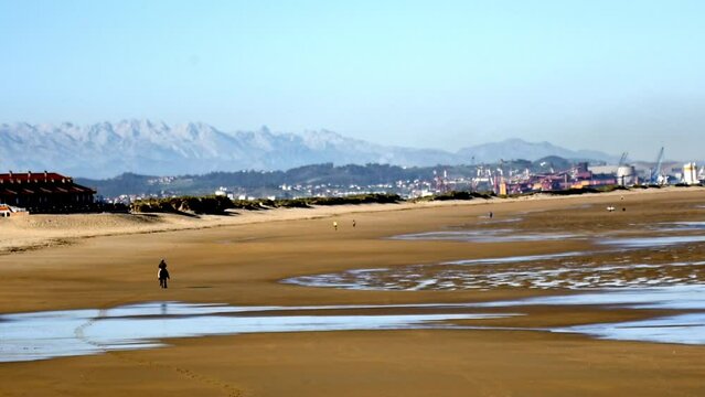 static wide angle shot of Somo beach in Santander where we see the mountains in the background in the foreground the beach with its puddles and a rider rides a horse on the beach on a sunny autumn day