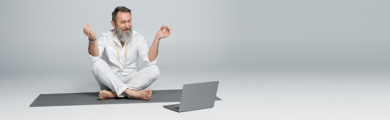 senior yoga coach showing chin mudra near laptop while sitting in easy pose on grey, banner.