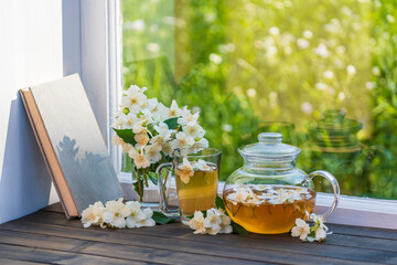 Hot herbal tea in glass teapot, cup and beautiful bouquet of jasmine flowers on windowsill at summer day near garden