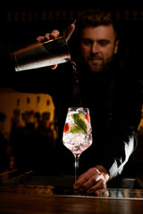 glass with ice cubes and bartender accurate pouring cocktail from shaker into it