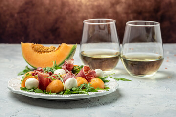 mediterranean salad with prosciutto or jamon, mozzarella and green basil leaves and Cantaloupe melon on white table, traditional Spanish and Italian appetizer served with wholemeal grissini, top view