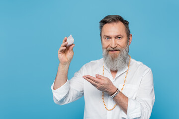 bearded master guru pointing at selenite crystal and looking at camera isolated on blue. - obrazy, fototapety, plakaty